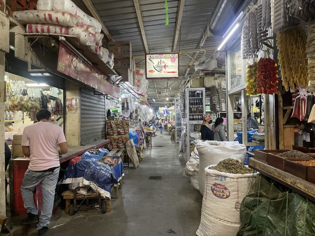 A Narrow Indoor Market Aisle Lined With Vendor Stalls Selling Spices, Dried Goods, And Sacks Of Grain, With Shoppers Walking Through.
