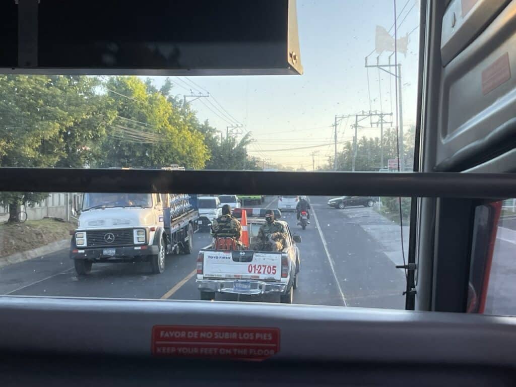 View Through A Bus Window Of A Busy Road, With A Delivery Truck, A Pickup Truck Carrying Two Soldiers, And A Motorcyclist Passing By.
