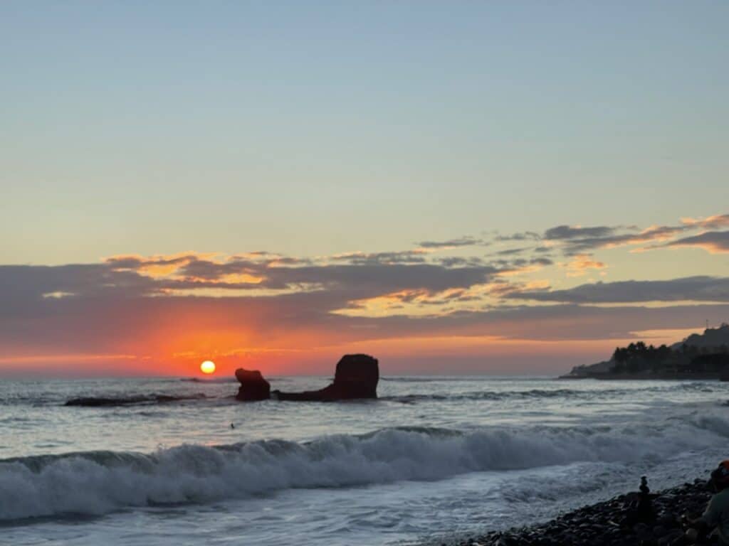 A Vivid Orange Sunset Over The Ocean With Tall Dark Rock Formations Rising Out Of The Surf Near The Shore.
