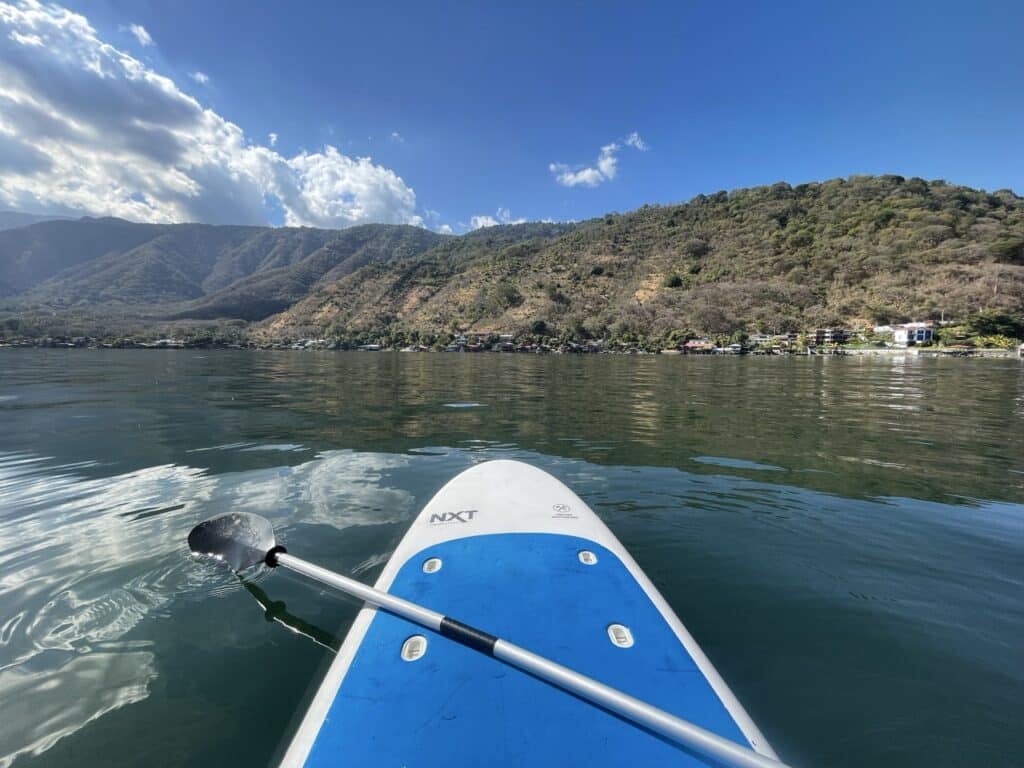 View From A Stand-Up Paddleboard Looking Across A Calm Lake Toward Green Hills And A Small Lakeside Town.
