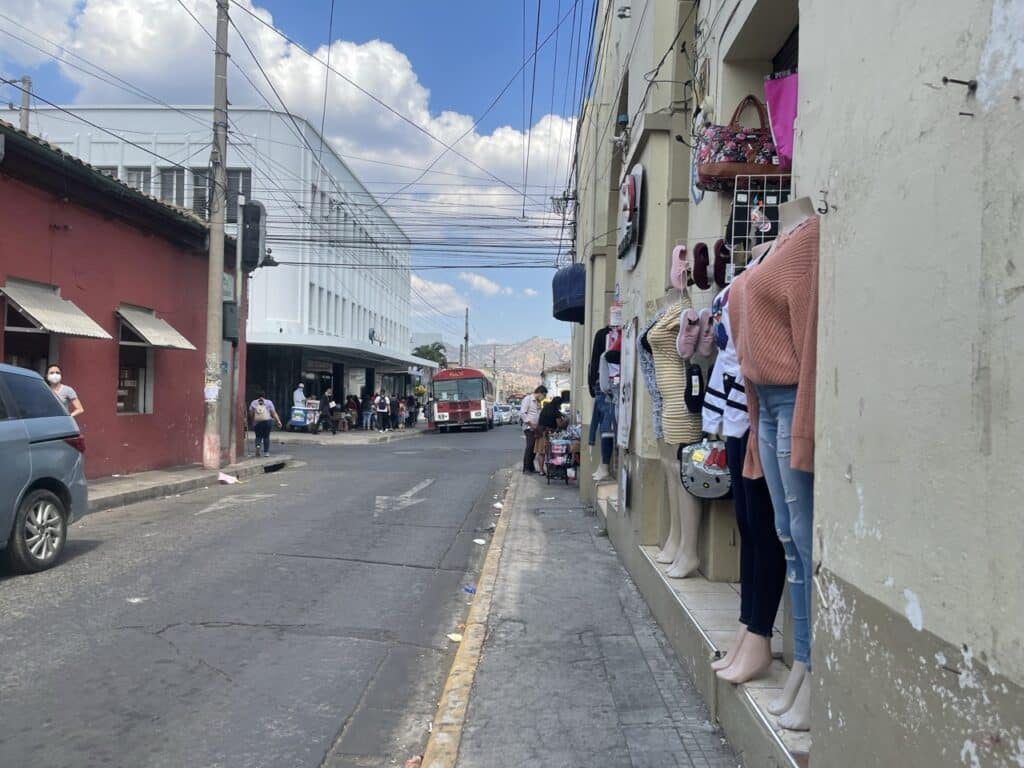A City Street Lined With Shops, Mannequins In A Clothing Store Window, And A Red Bus Driving Past Under A Partly Cloudy Sky.