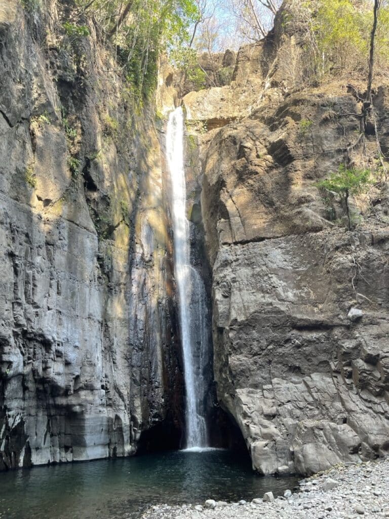 A Tall Waterfall Pouring Down A Rocky Cliff Into A Pool Surrounded By Stone Walls And Greenery.